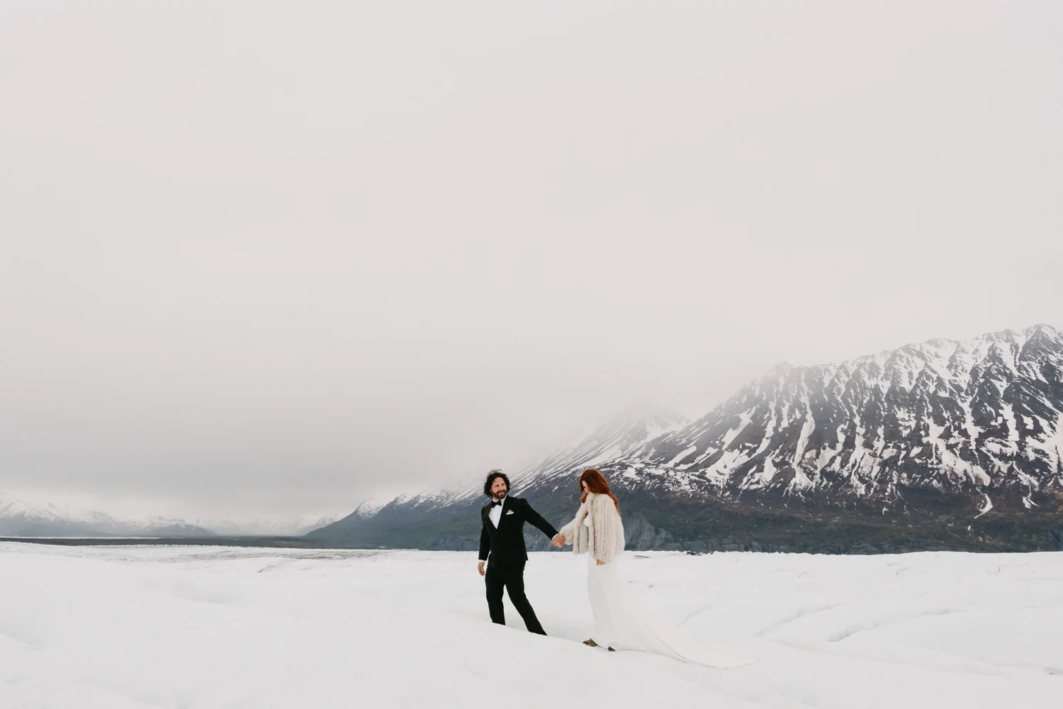 A bride and groom holding hands in a snowy landscape with mountains in the background.