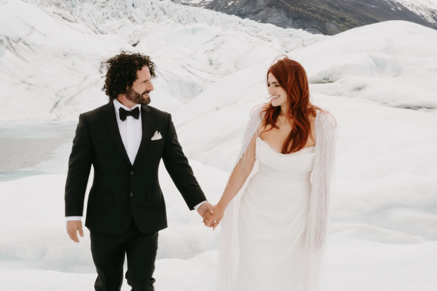 A bride and groom stand hand in hand on a snowy landscape, surrounded by glaciers with mountains in the background. The groom is dressed in a black tuxedo with a bow tie, and the bride is wearing a white wedding gown.