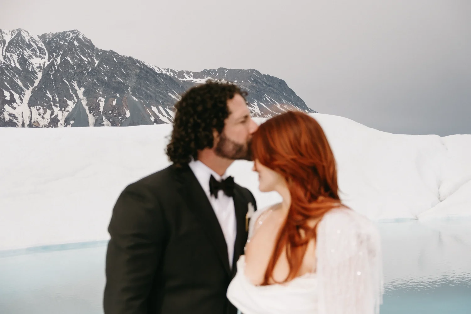 A man in a tuxedo with a bowtie kissing a red-haired woman in a white dress near a snowy landscape with a mountain in the background.