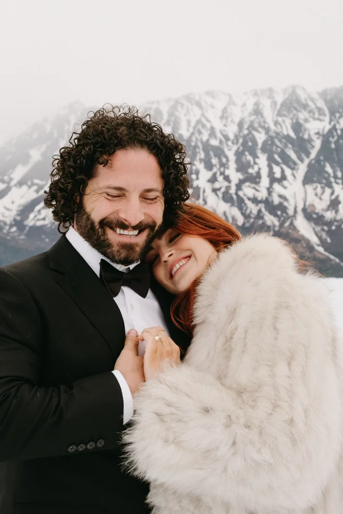 A smiling couple embracing outdoors, with a snowy mountain range in the background.