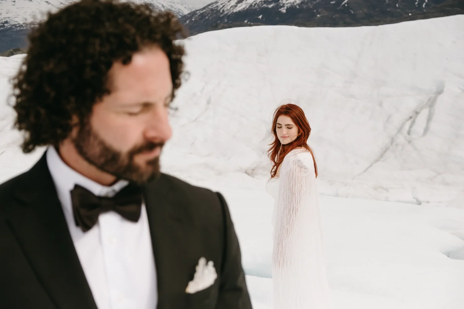 A man in a tuxedo with a bow tie in the foreground, blurred, with a woman in a white dress with long sleeves, standing in the background in a snowy landscape with snow-covered mountains.