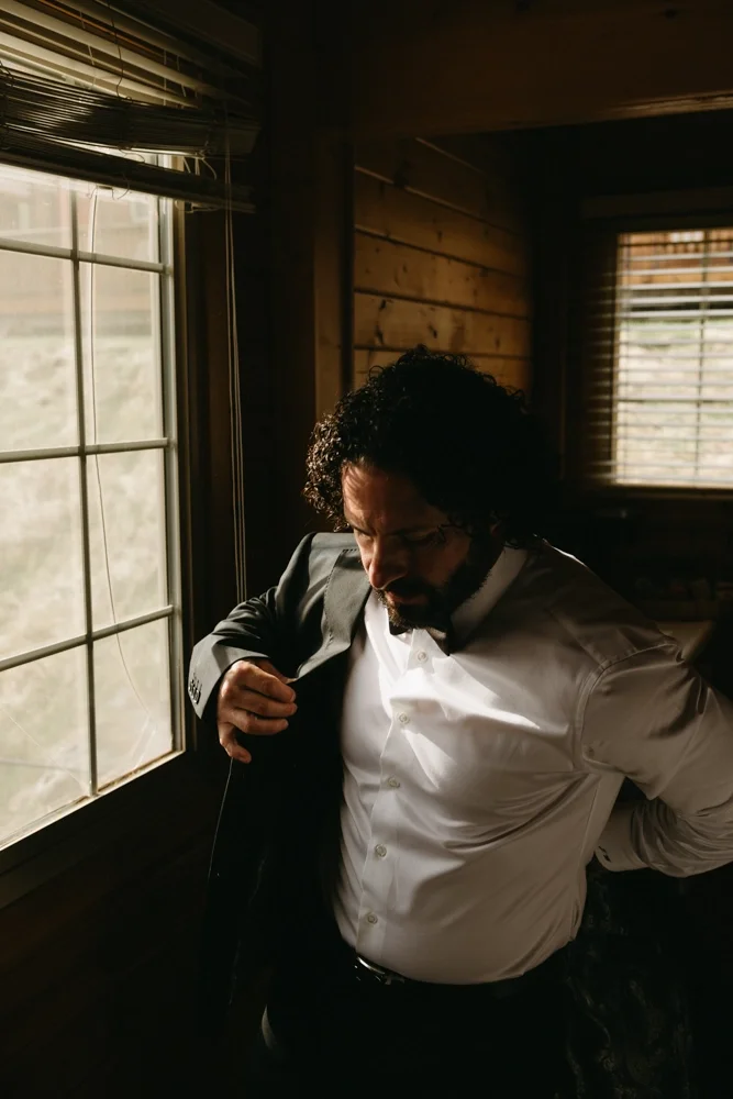 A man with curly hair and a beard adjusting his suit jacket in a wooden room, standing by a window with blinds, with natural light illuminating his face.