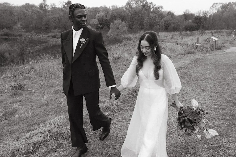 A bride and groom in wedding attire hold hands while walking through a field at the Trinity River Audubon Center.