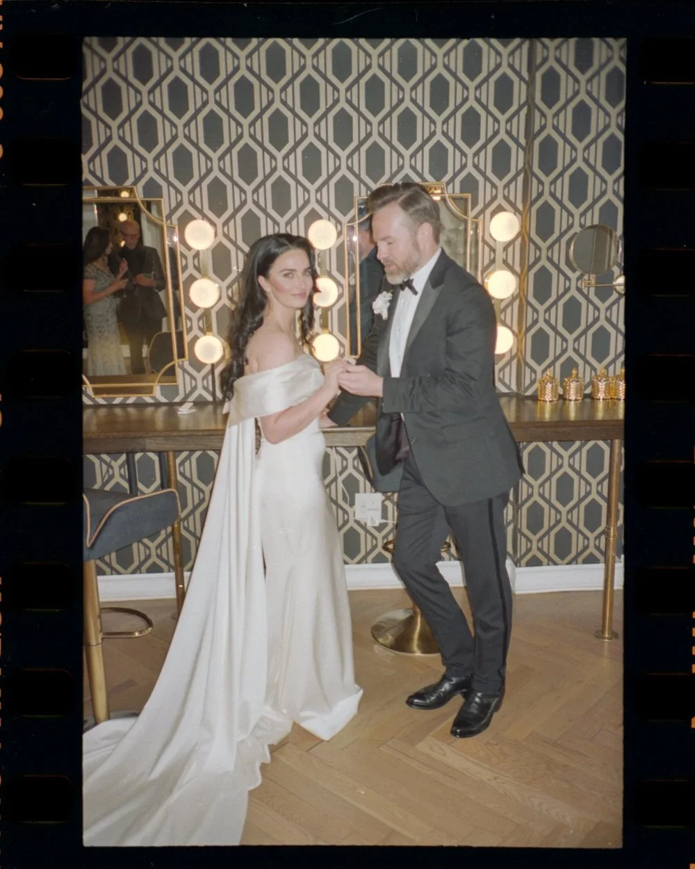 A bride in an off-the-shoulder gown and groom in a tuxedo hold hands before a vanity mirror, captured by a Dallas wedding photographer.