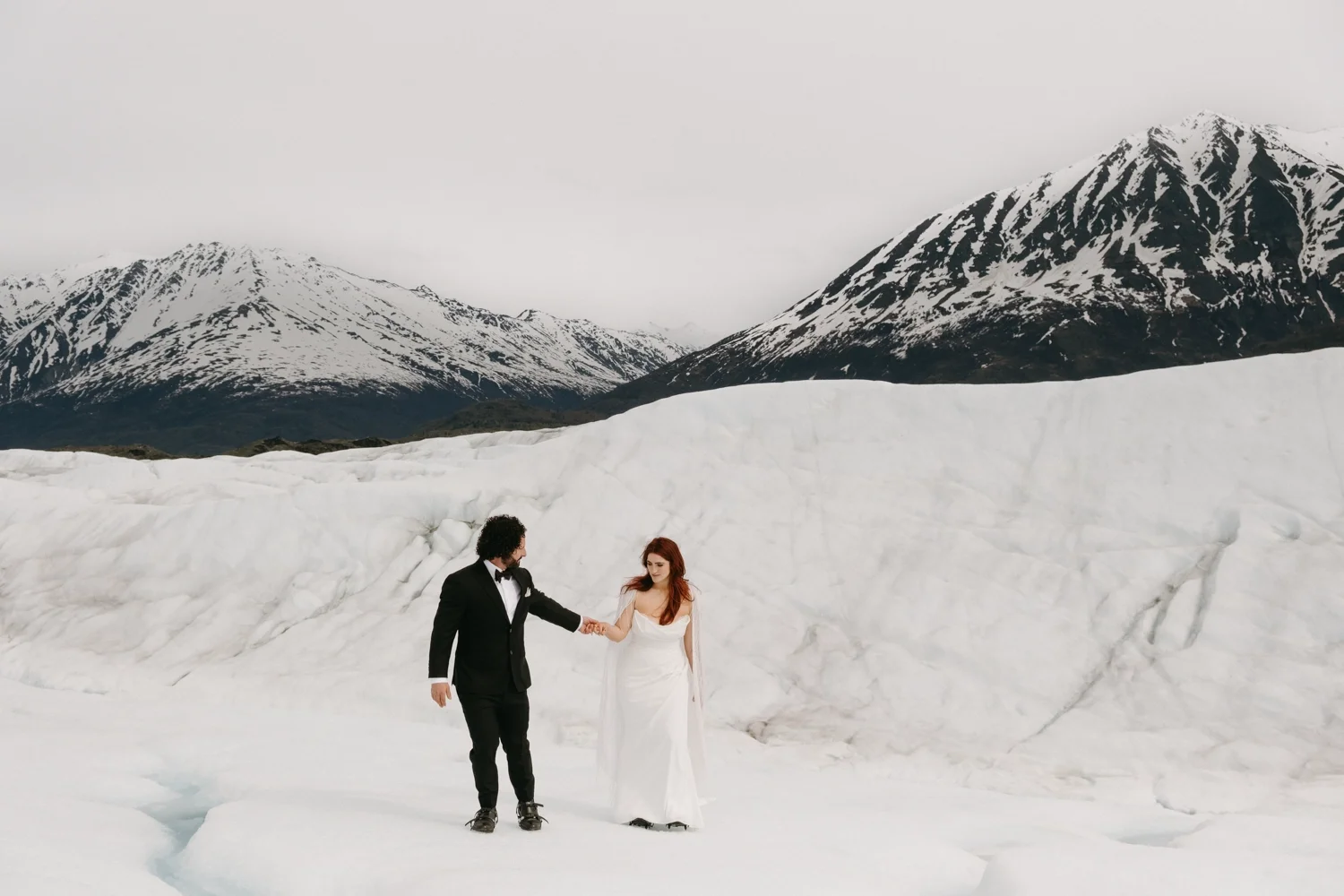 A bride and groom holding hands on a snowy glacier in a mountainous, snowy landscape, with snow-capped mountains in the background.