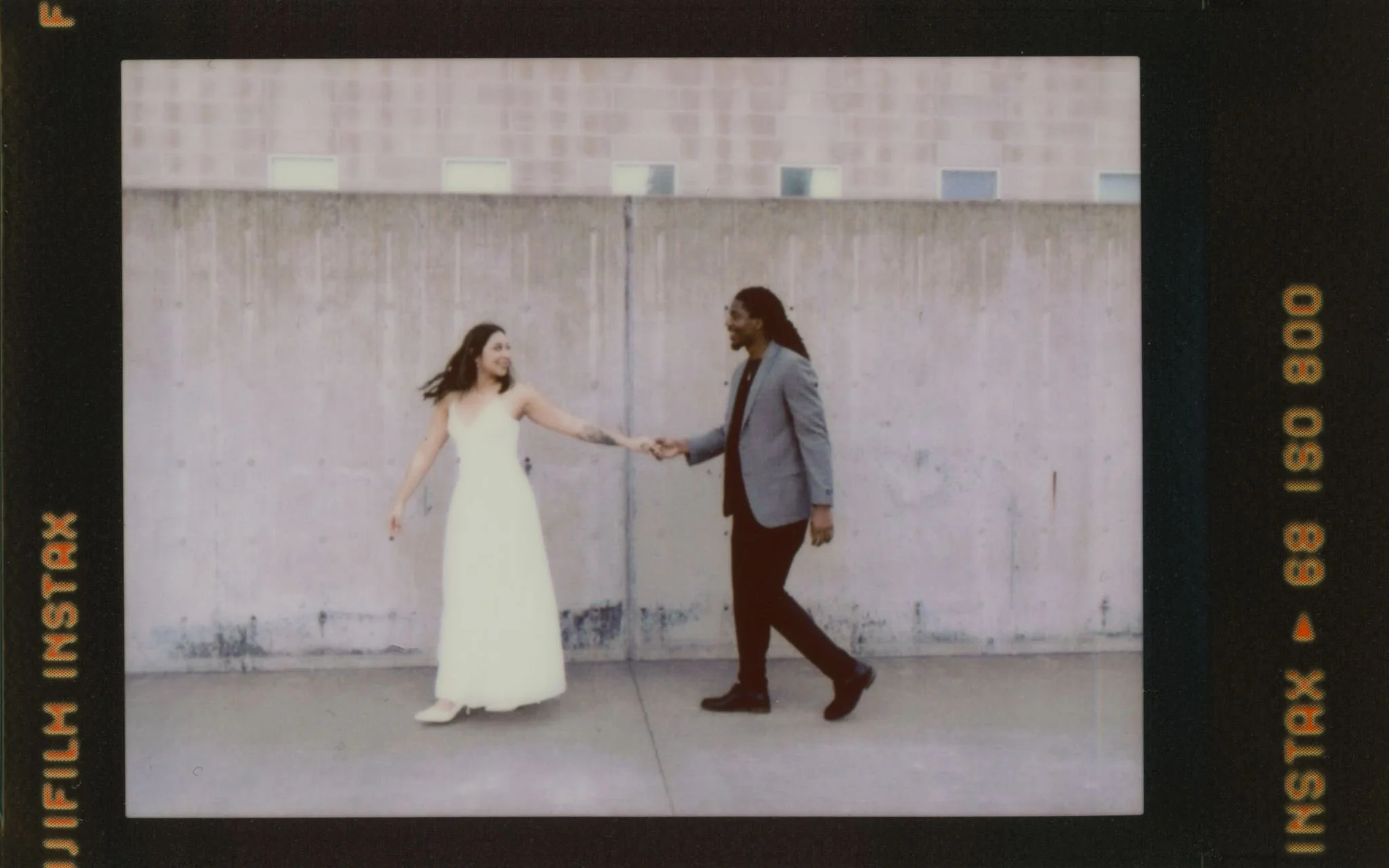 A woman in a white dress and a man in a blazer hold hands while walking in front of a concrete wall, captured on Instax Mini.