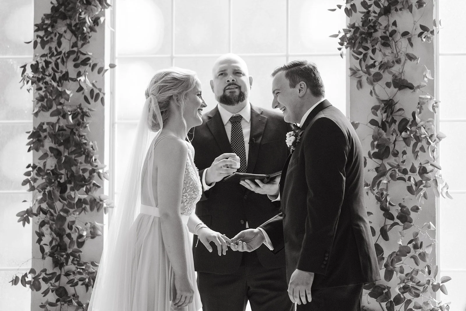 A bride and groom exchange rings during their wedding ceremony at The Emerson, officiated by a man standing between them.
