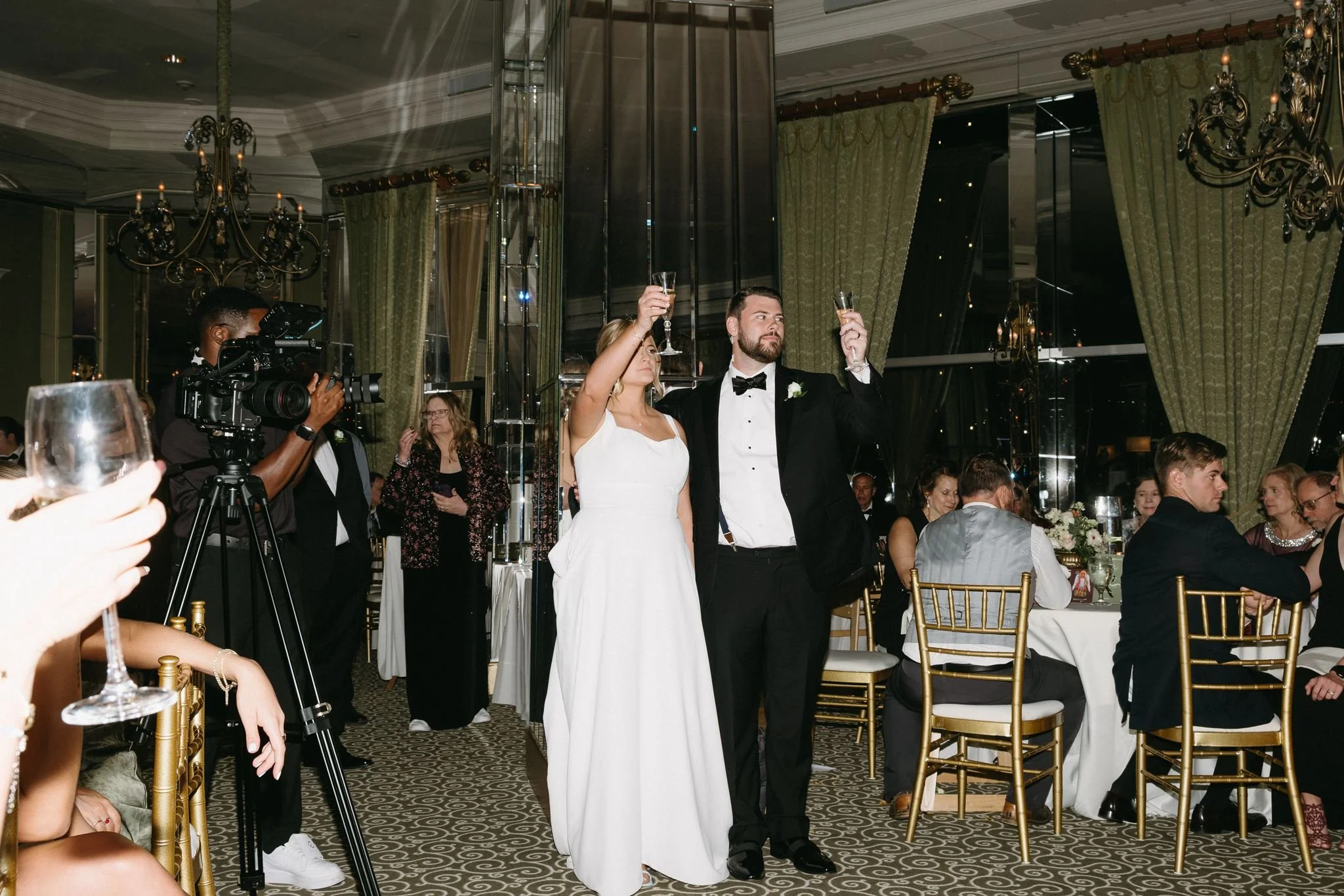 A bride and groom stand in the City Club of Fort Worth ballroom during a wedding toast, surrounded by guests and a videographer.