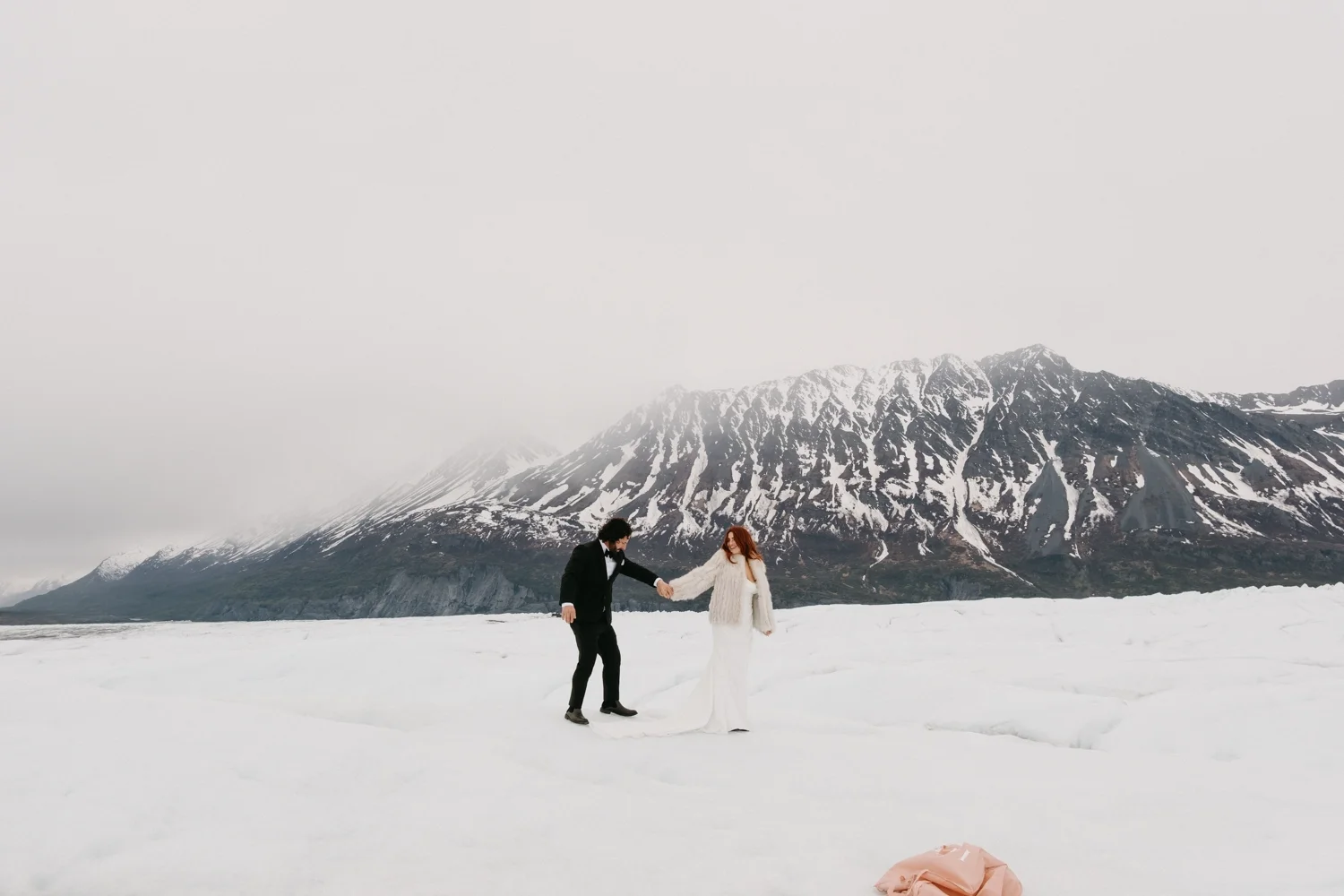 A couple in wedding attire holding hands in a snowy landscape with mountains in the background.