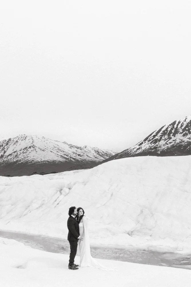 A couple dressed in wedding attire standing on snowy terrain near a large ice formation with snow-covered mountains in the background.