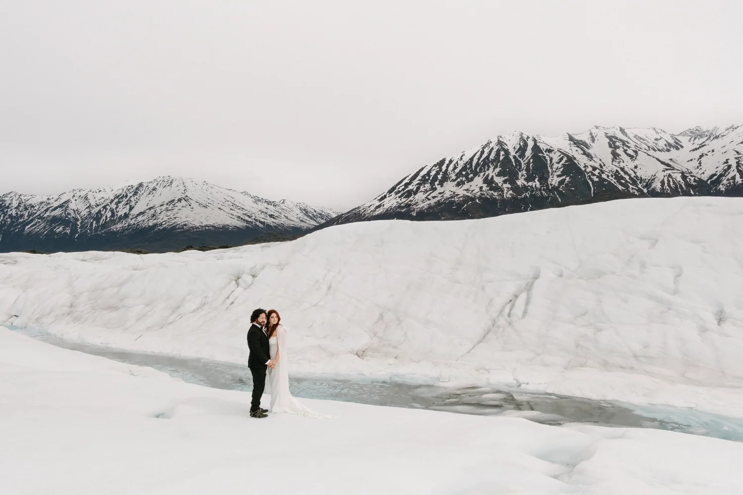 A couple dressed in wedding attire stands on snow near a partially frozen body of water, with snow-capped mountains in the background.