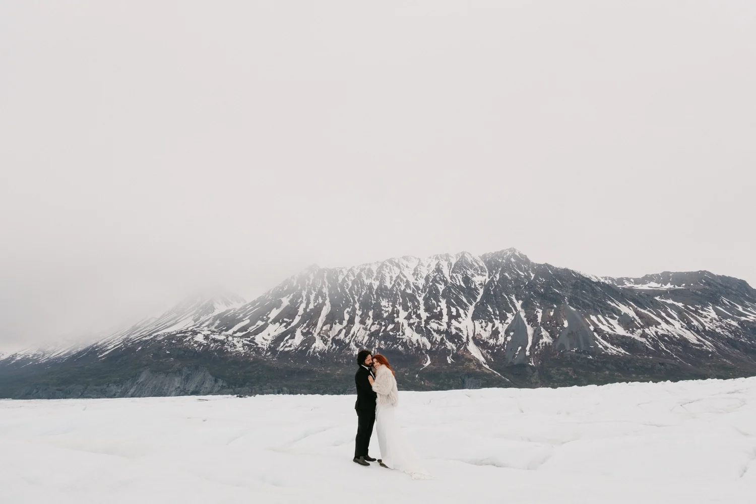 Couple in wedding attire standing on snow-covered landscape with snow-capped mountain in the background.