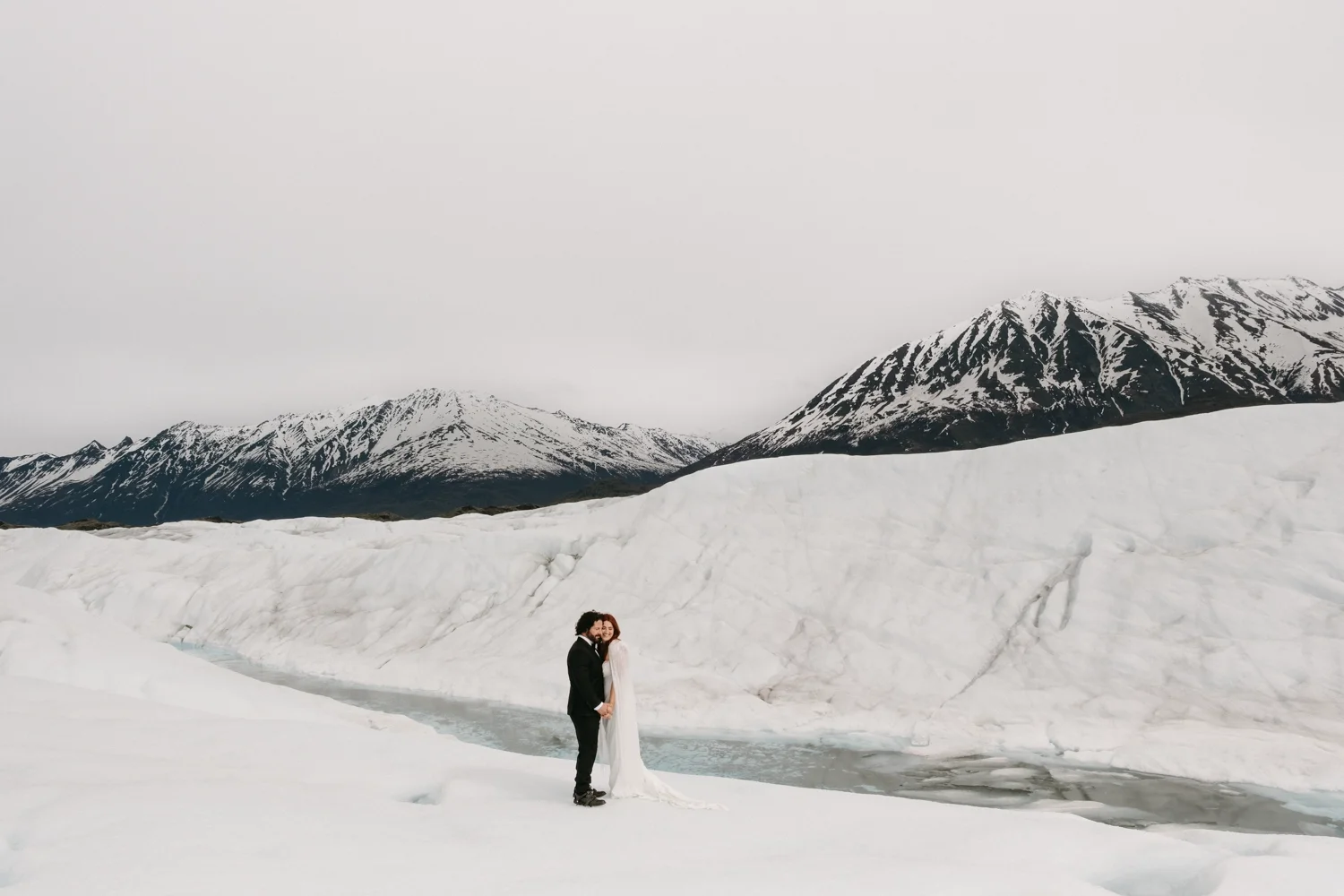 Alaska Glacier Wedding | A couple dressed in wedding attire holding hands and smiling in a snow-covered landscape with mountains in the background.