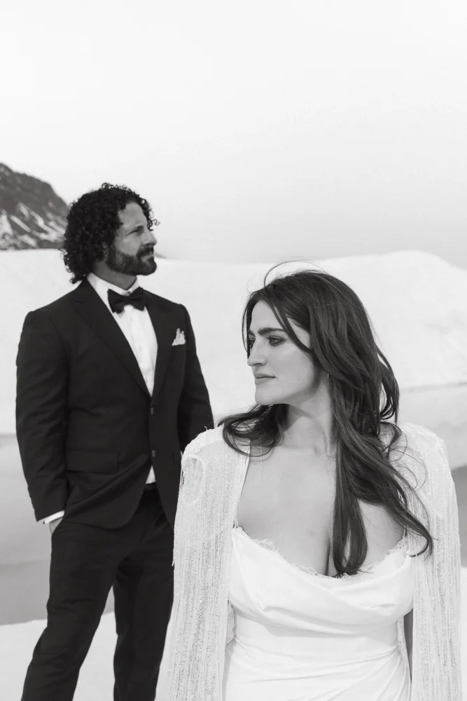 A man in a tuxedo and a woman in a wedding dress are outdoors on a snowy landscape, with a mountain in the background.