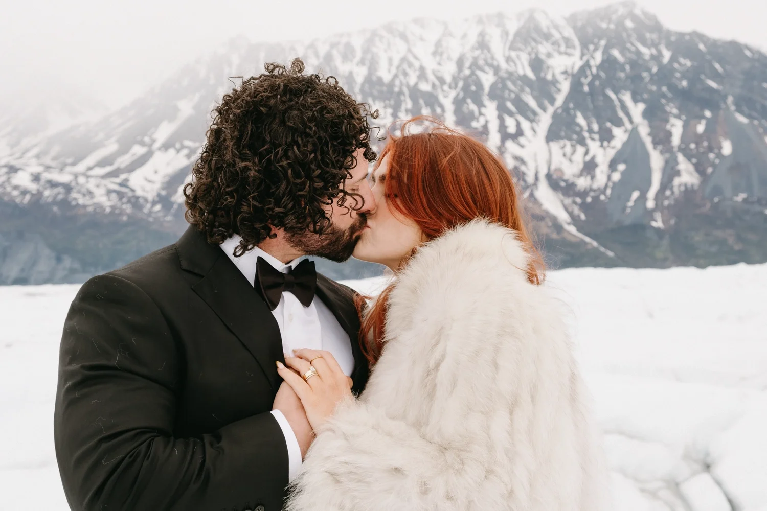 A man and woman kissing outdoors in a snowy mountain landscape, the man in a black tuxedo and the woman in a white fur coat.