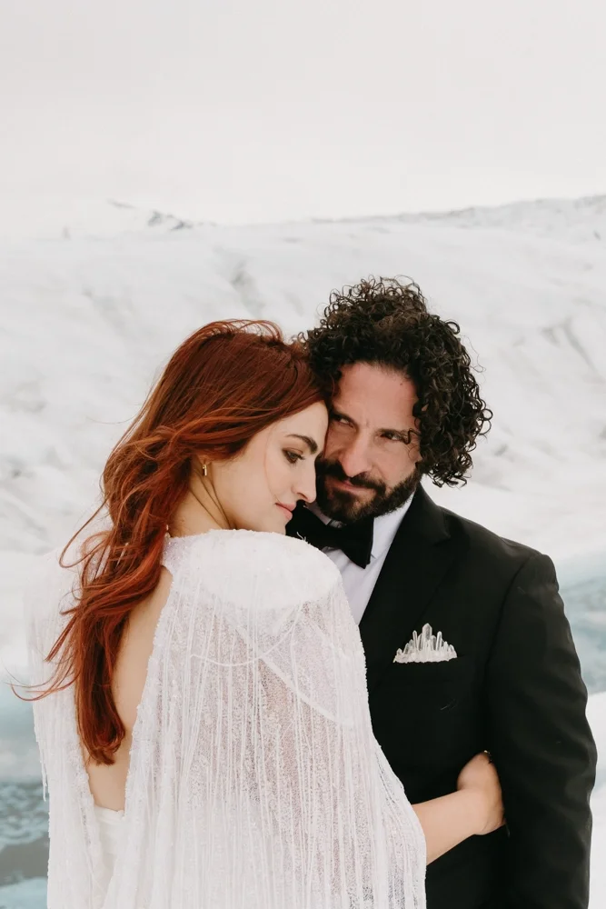 A couple dressed in wedding attire embracing on a snowy landscape.