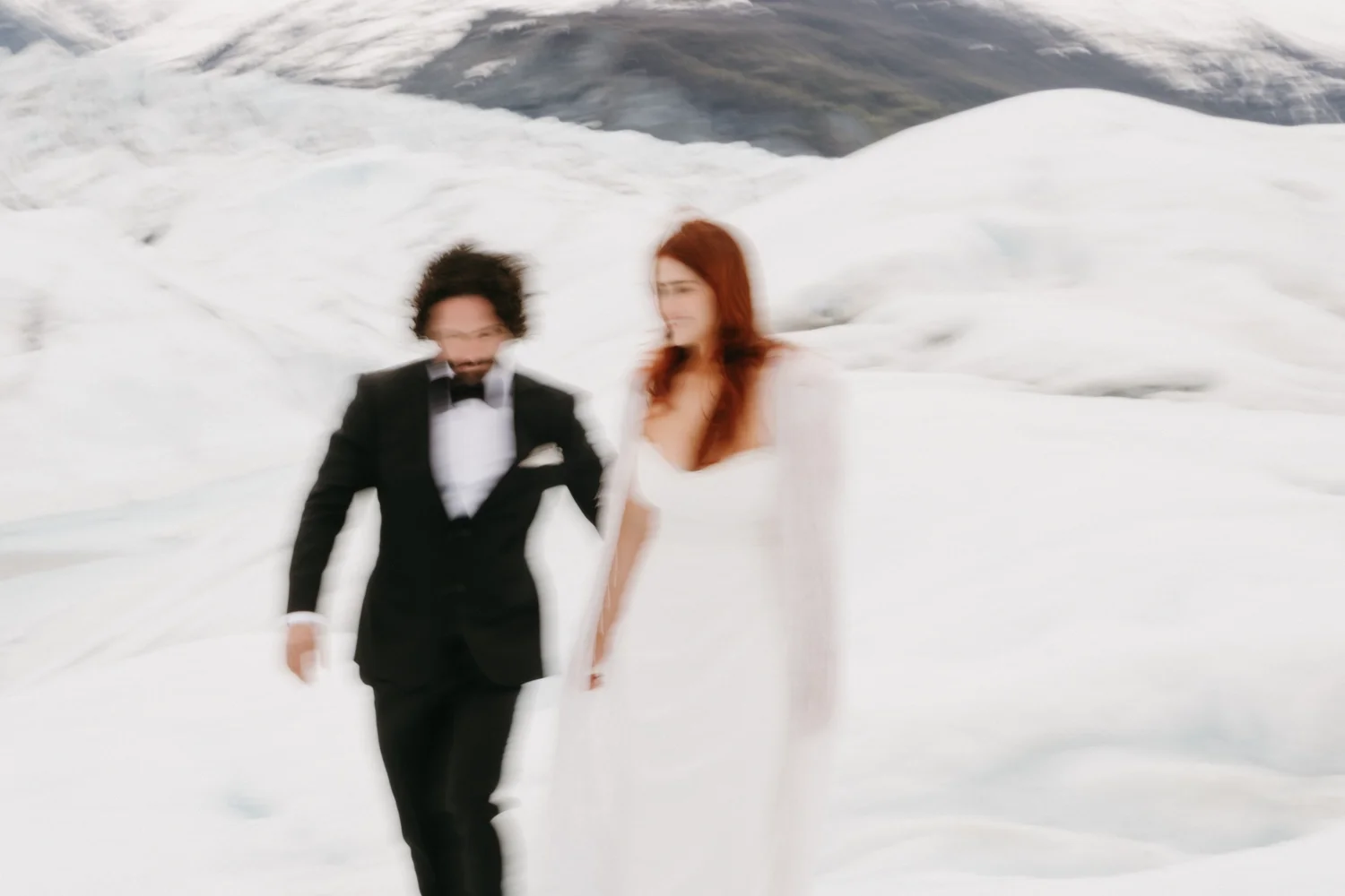 A bride and groom walking hand-in-hand on snowy terrain with ice and snow formations in the background.