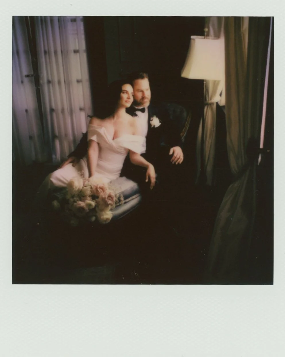 A bride in a white gown and a groom in a tuxedo sitting together with a bouquet of flowers in a color i-type Polaroid photo.