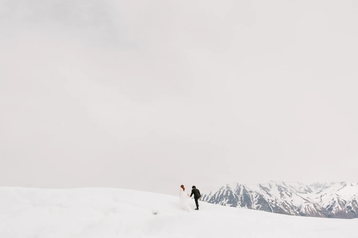A couple dressed in wedding attire walking hand in hand on snow-covered ground with snow-capped mountains in the background.