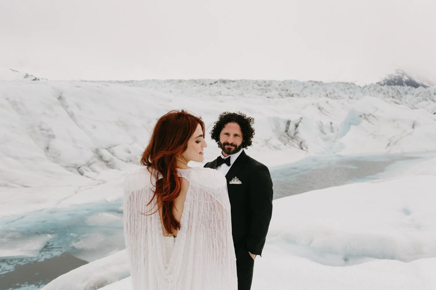 A couple dressed in wedding attire standing on snow and ice in a glacial landscape, with a woman in a white dress and a man in a black tuxedo.