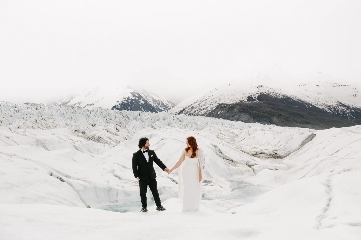 A bride and groom holding hands in a snowy glacier landscape with mountains in the background.