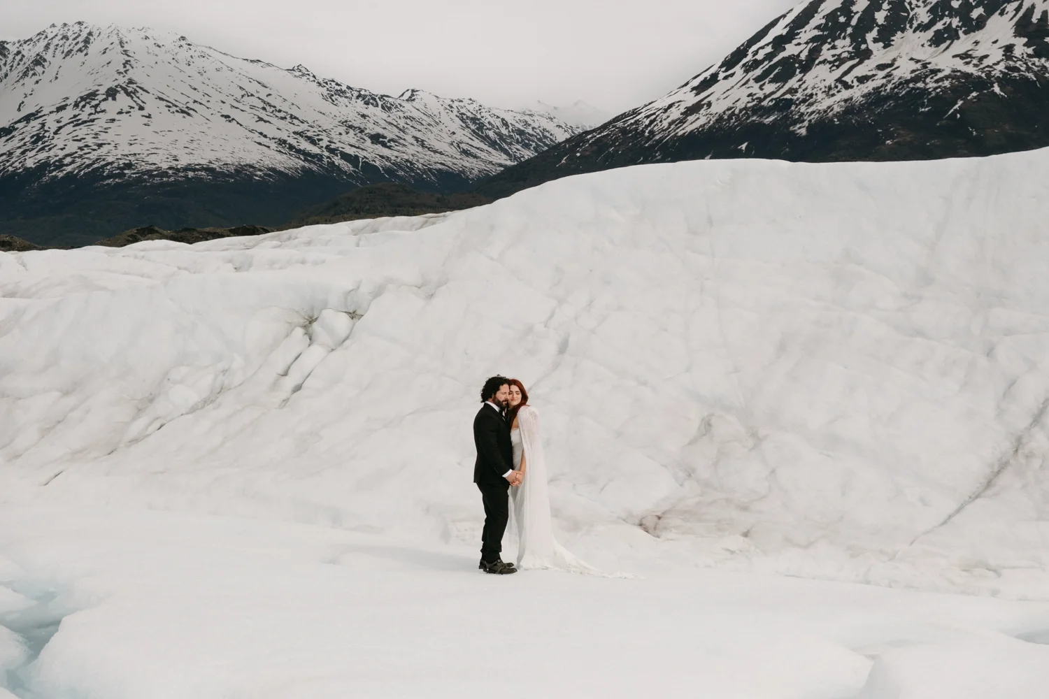 A bride and groom in wedding attire standing on ice in a snowy mountainous landscape.