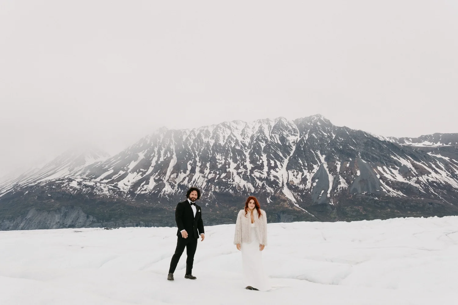 A man in formal tuxedo and a woman in a wedding dress standing on snow with a mountain background.