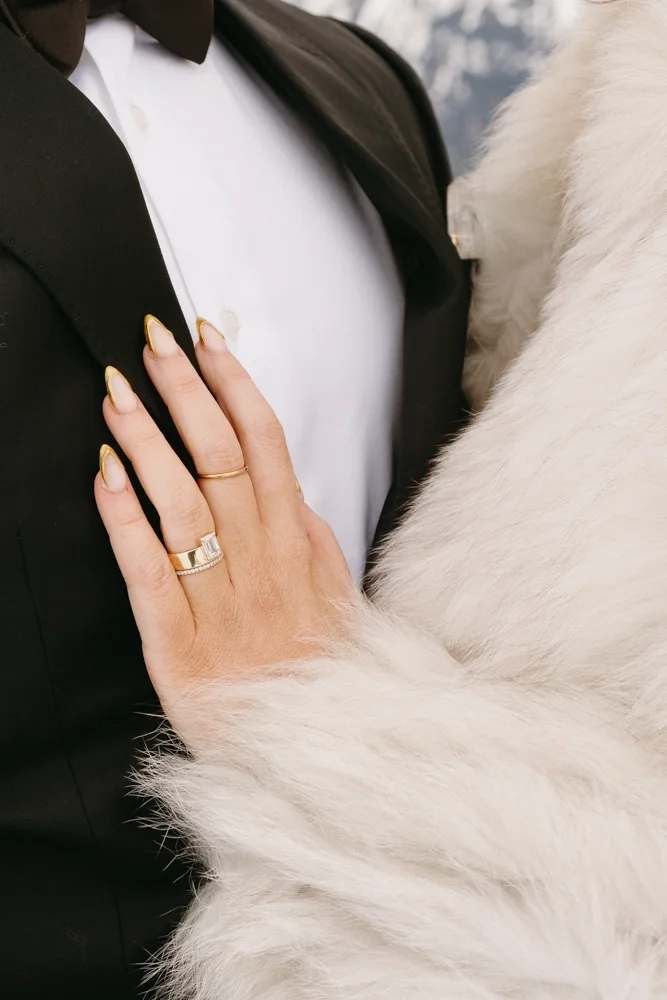 A close-up of a woman's hand resting on a person's chest, wearing rings and gold-accented nails, with a white fur coat and black suit jacket visible.