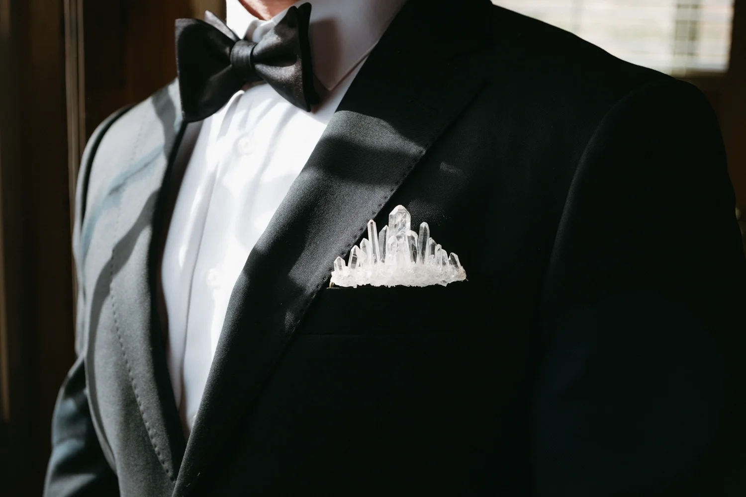 A man in a tuxedo with a black bow tie and a crystal-like lapel pin on the left side of the jacket.