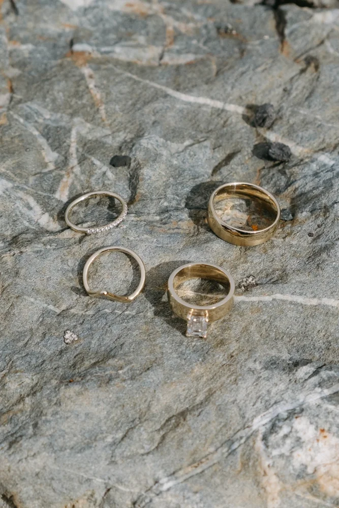 Four rings placed on a rough, gray stone surface. One ring has small diamonds, another appears to be plain gold, and the third has a prominent square-cut gemstone.