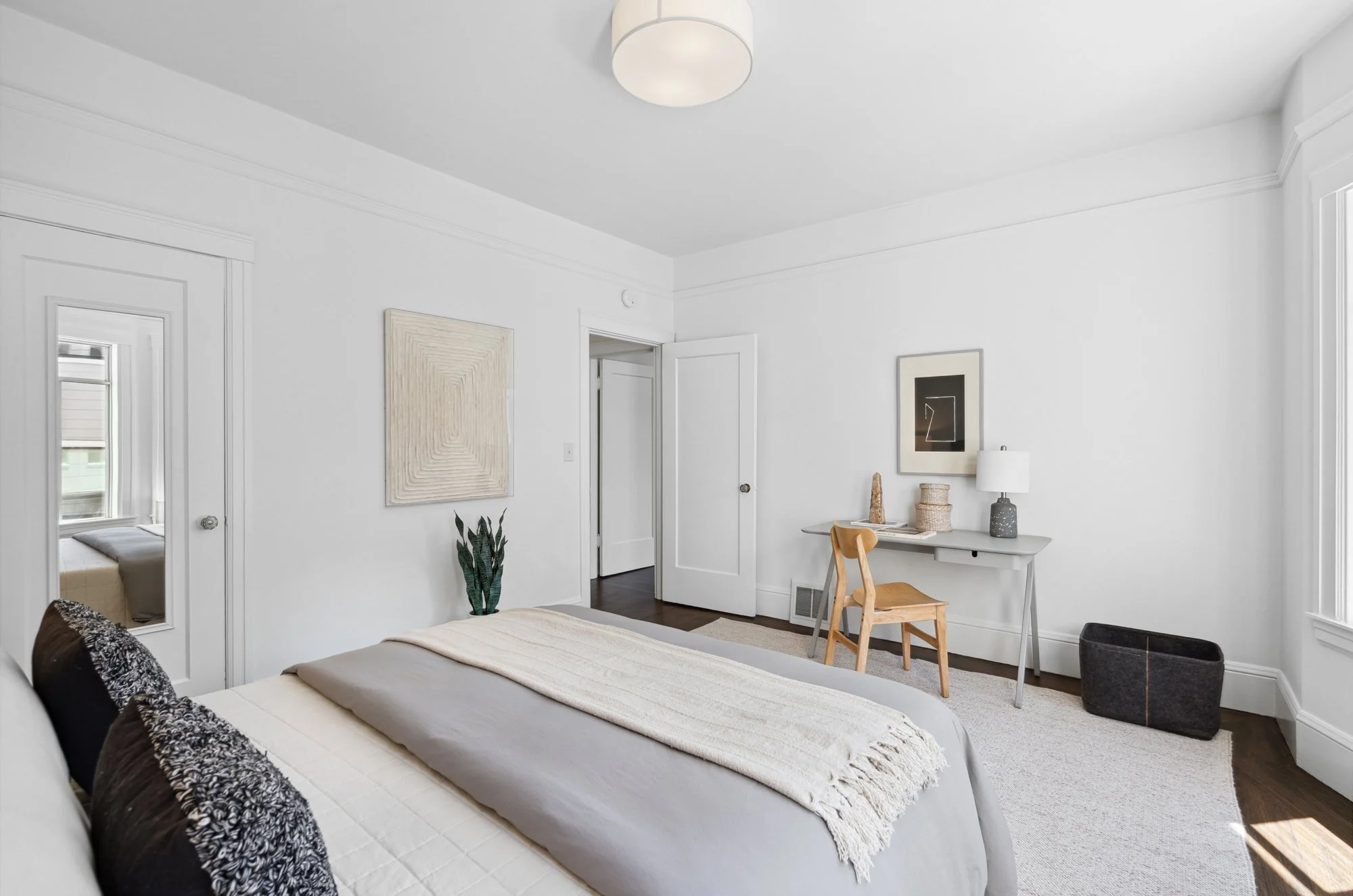 A bright, white bedroom with a bed in the foreground, a desk and chair to the right wall, and wall decor including framed art, a table lamp, and a potted plant. There are windows on the right and in the reflection of a mirror on the left, with white 