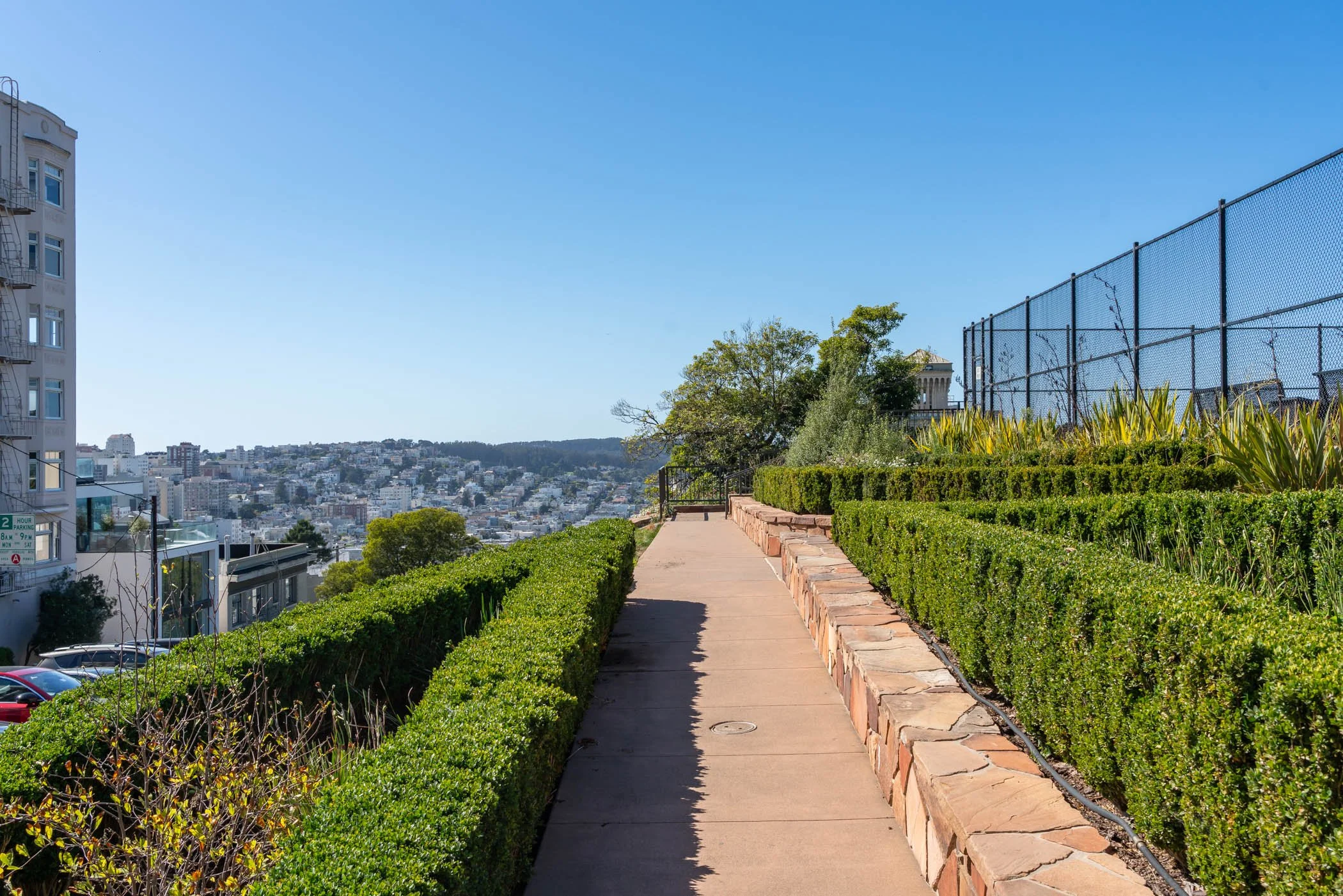 A sidewalk with a low brick wall on the right and neatly trimmed bushes, leading to a cityscape view with buildings and a hill in the background, under a clear blue sky.
