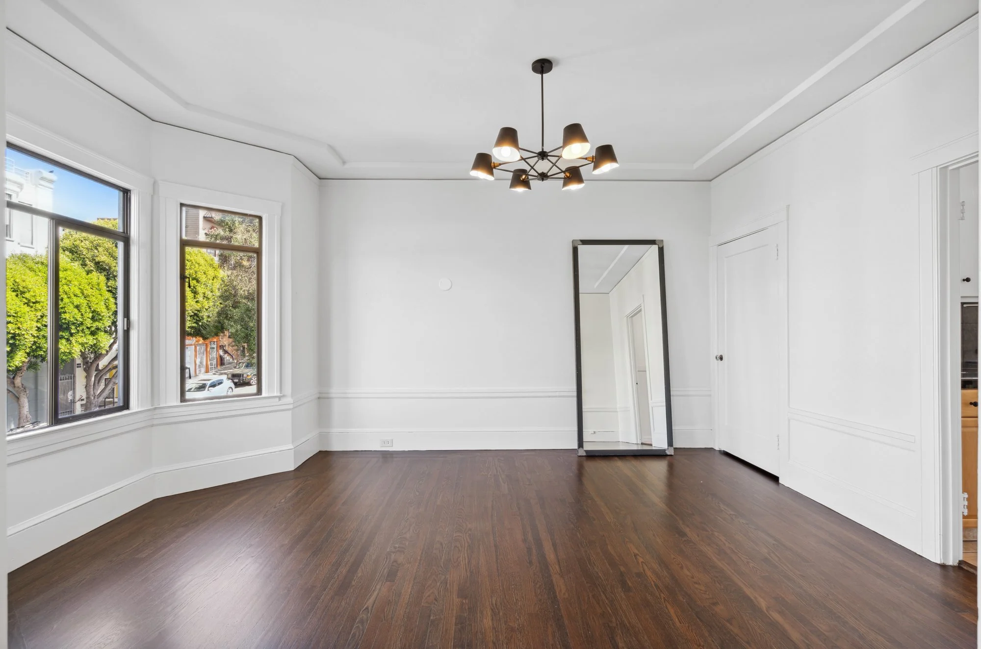 Empty room with large windows, white walls, a ceiling light fixture, hardwood floors, and a full-length mirror leaning against the wall.
