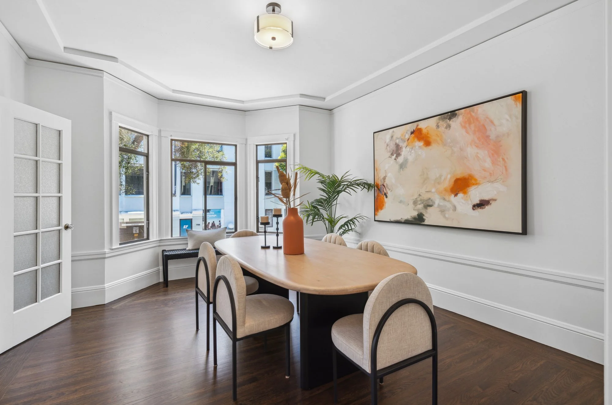 Bright dining room with a wooden table, beige upholstered chairs, large windows, a potted plant, a large abstract painting, and a ceiling light fixture.