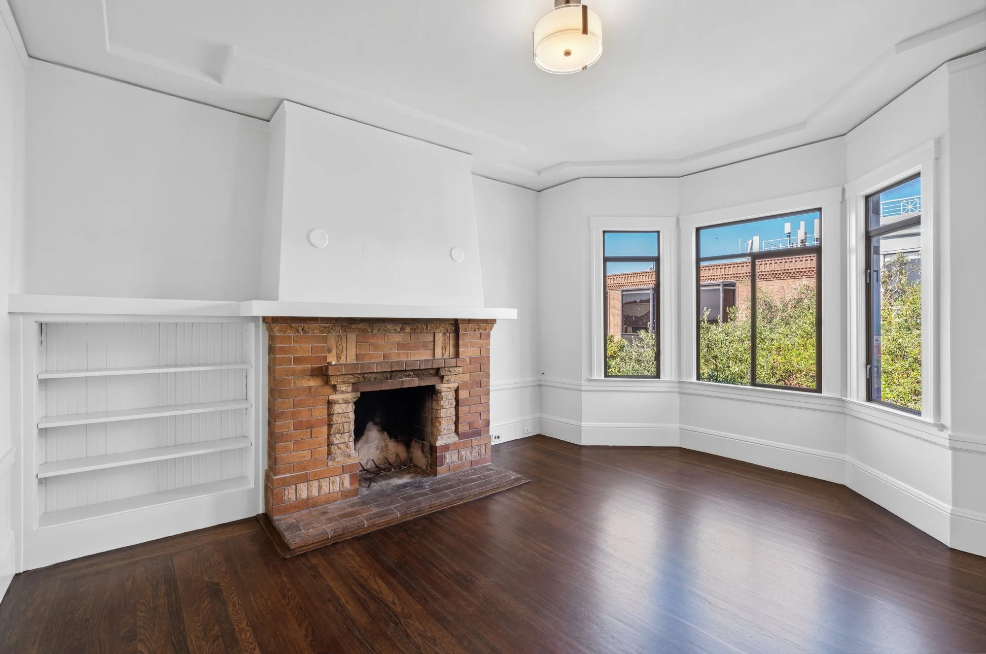 Living room with white walls, hardwood floor, a brick fireplace, and three large windows showing a building and trees outside.