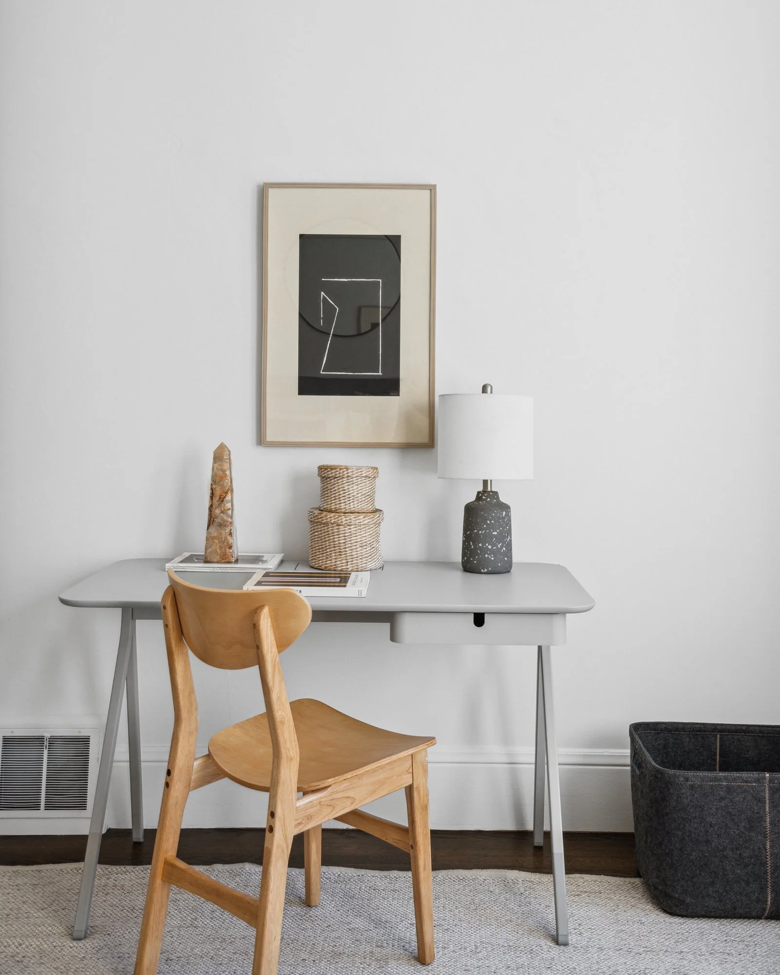 A simple room corner with a white wall, a framed black and white abstract art piece, a white table, a wooden chair, a decorative stone sculpture, a woven basket, a white table lamp, and a black fabric storage bin.