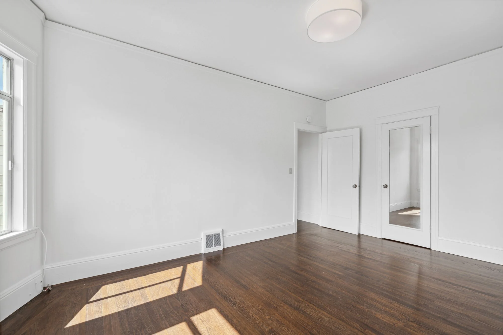 Empty room with white walls, hardwood floors, a window, and a mirror on the door.