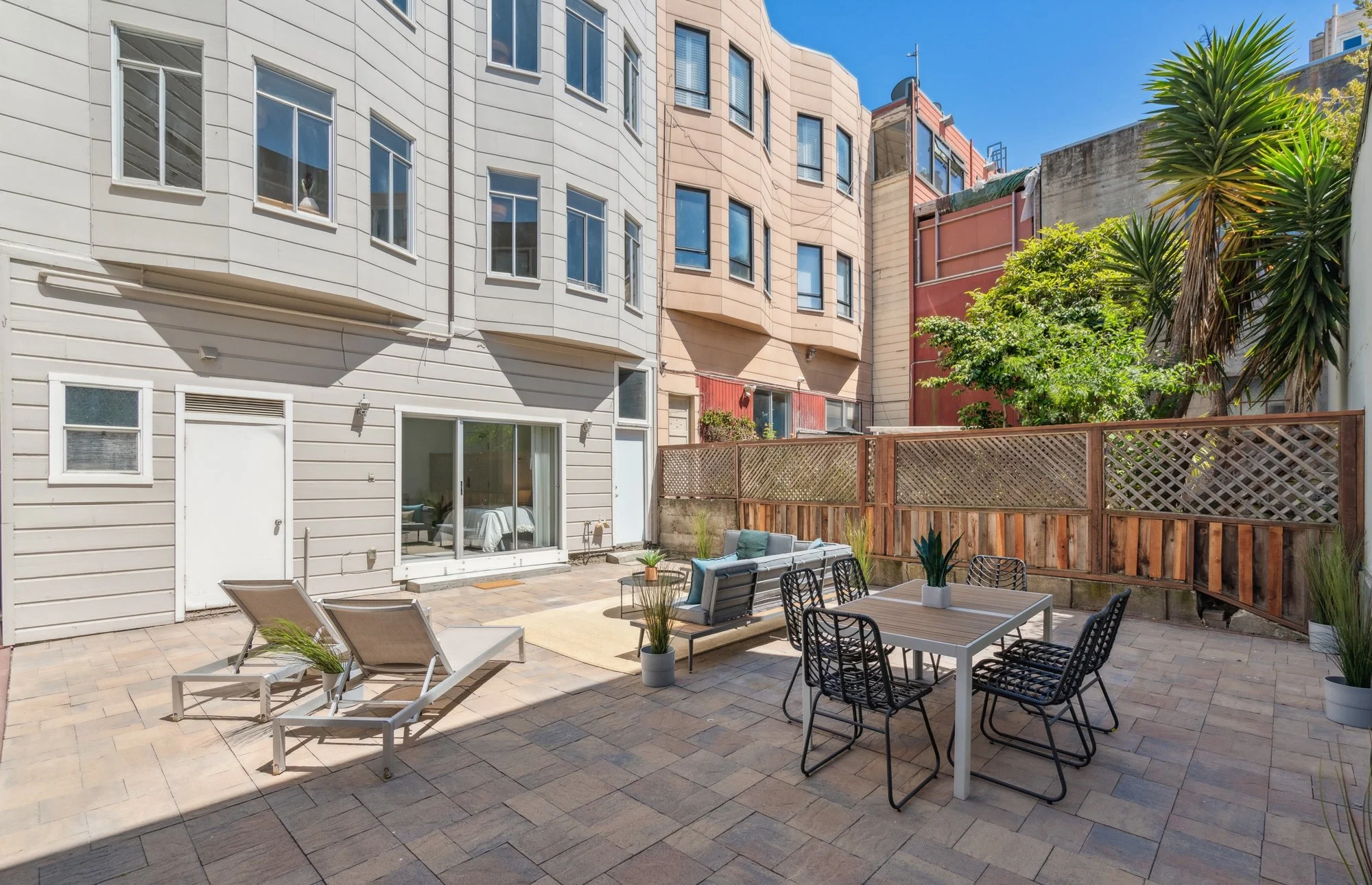 Outdoor patio with seating area, including lounge chairs, a sofa, and a dining table with chairs, surrounded by a wooden fence and greenery, with multi-story residential buildings in the background under a blue sky.