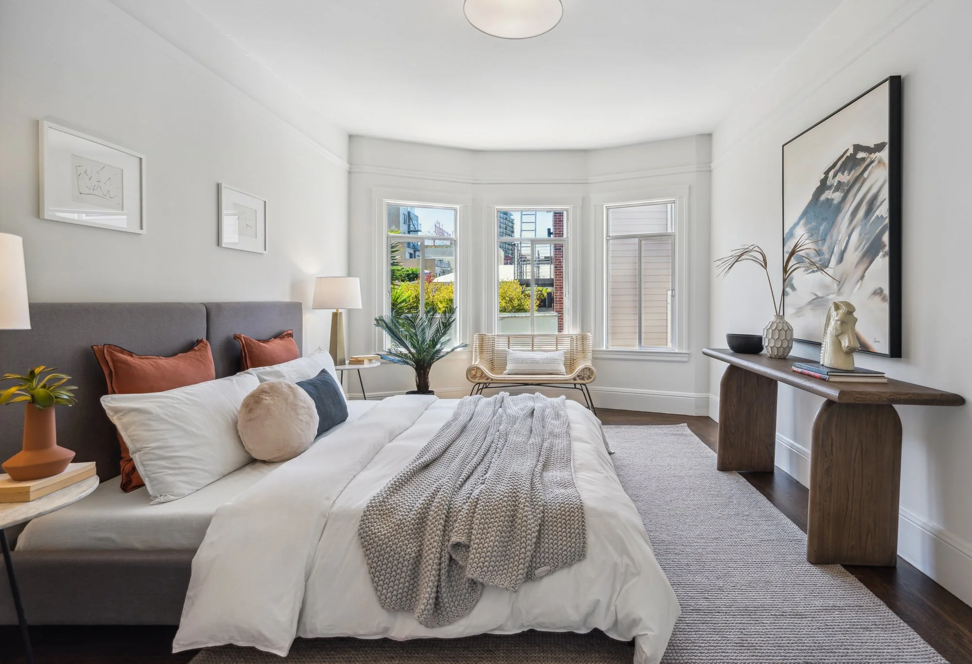 Bright bedroom with large bay windows, white walls, and artwork. Features a white bed with pillows and a blanket, a wooden side table with a lamp, a wicker chair, and a console table with vases and a sculpture.