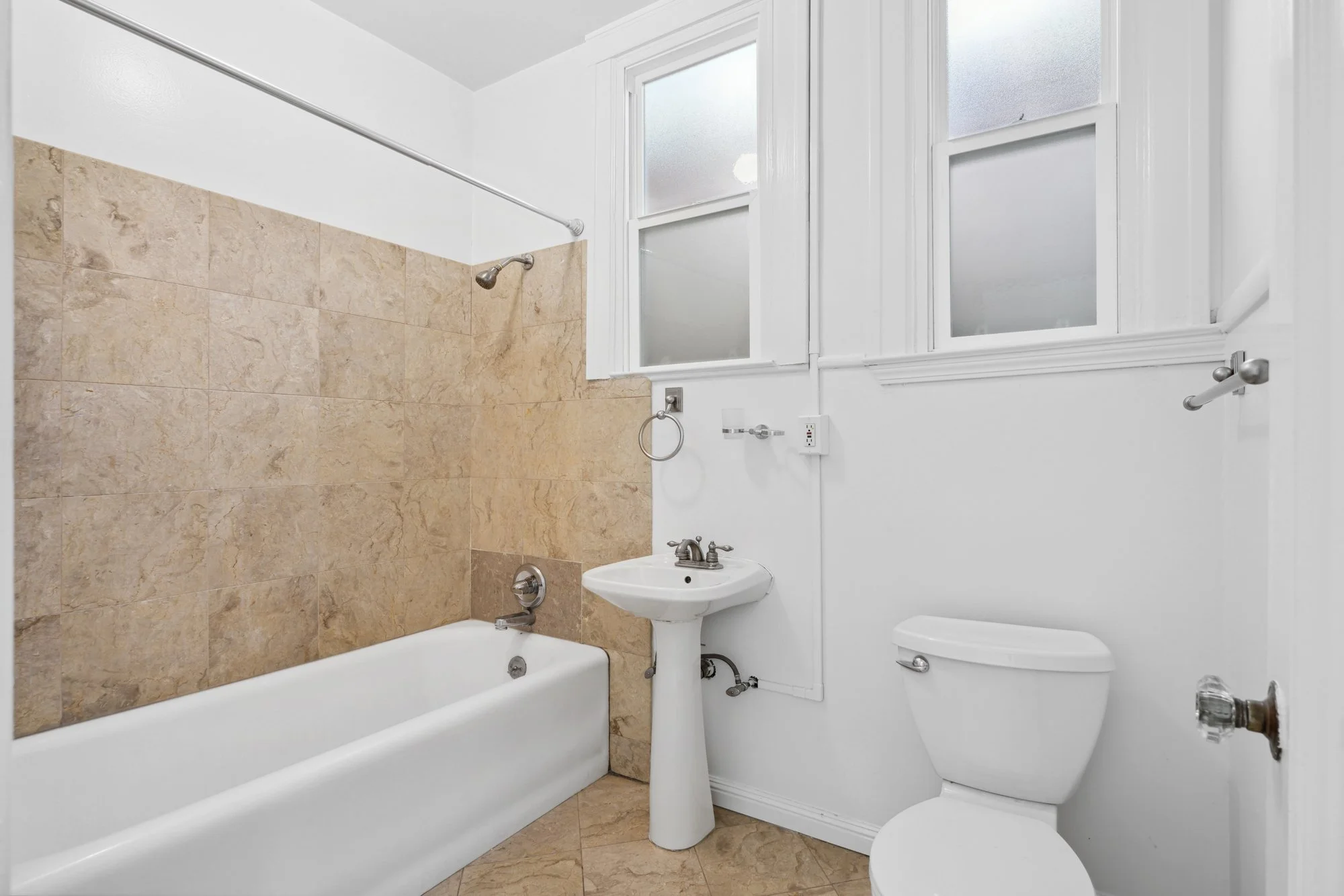 Bathroom with bathtub, shower, small sink, toilet, and large frosted windows, with white walls and beige tiled accents.