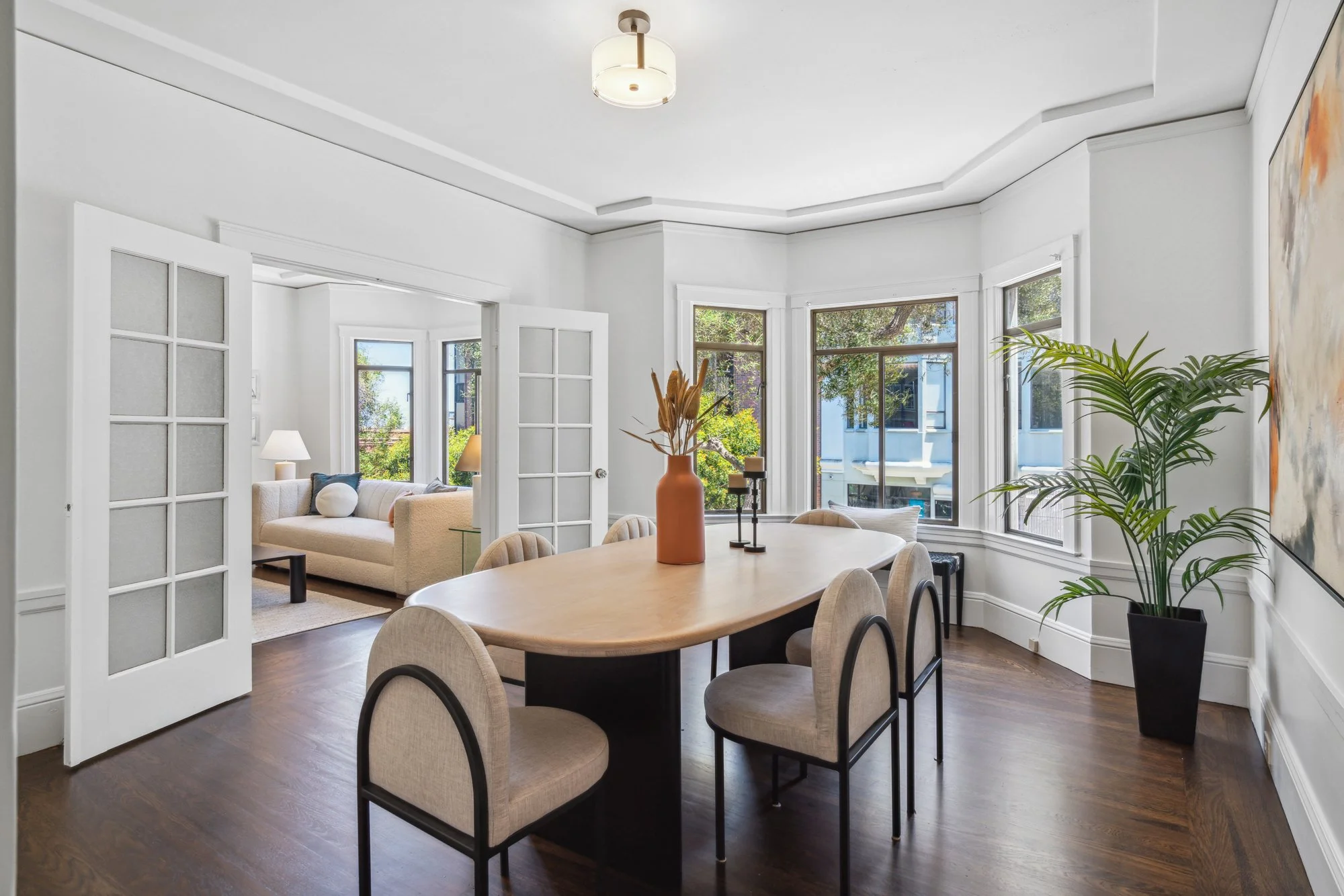 Bright, modern living room with large windows, white walls, wooden flooring, a beige sofa with cushions, a white side table with lamps, a round wooden dining table with beige chairs, decorative plants, and abstract wall art.