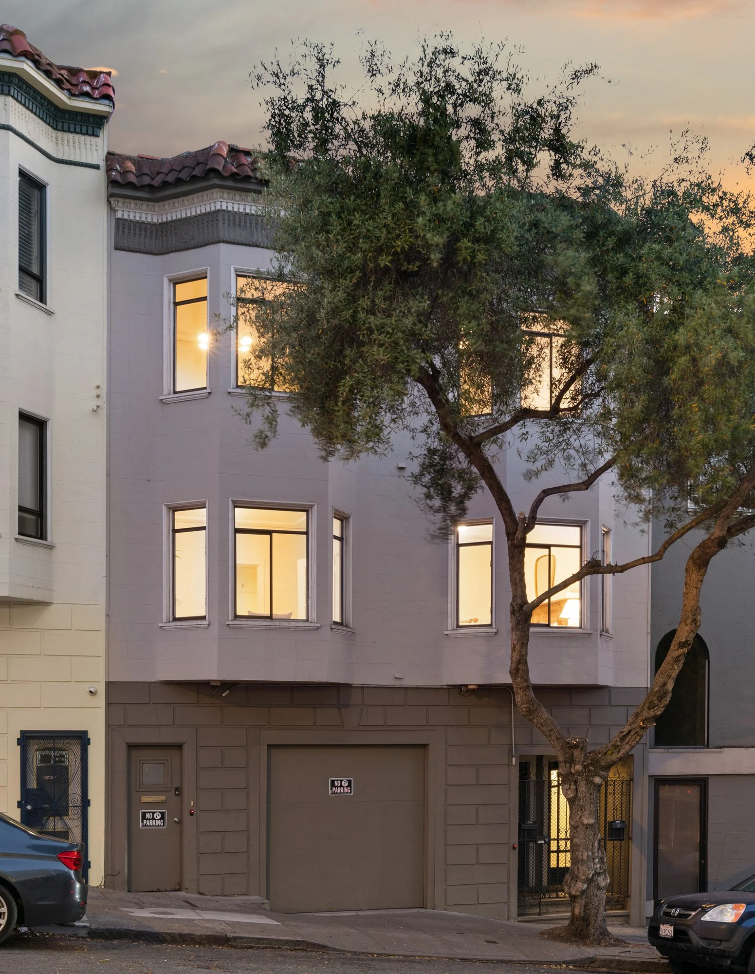 A three-story building with a gray exterior and large windows with glow from interior lights. The building has a tiled roof and decorative trim. There is a tall tree with a twisted trunk and spreading branches in front. Cars are parked along the stre