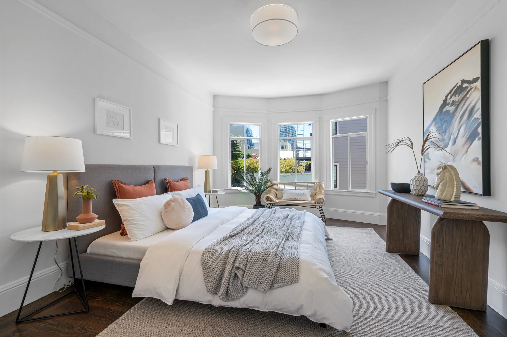 Bright bedroom with white walls, large windows, a bed with gray headboard, white and rust-colored pillows, and a throw blanket. Nightstands with lamps and decor, a rattan chair, art on the walls, and a wooden console table with plants and sculptures.