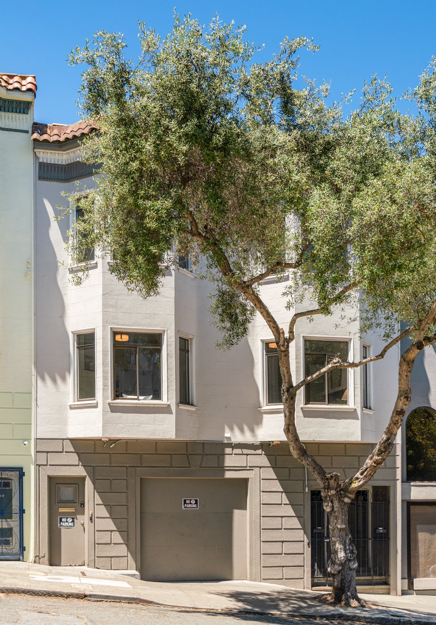 Front of a white building with three windows, garage, and two 'No Parking' signs, a tree with sparse leaves in front, and a blue sky above.