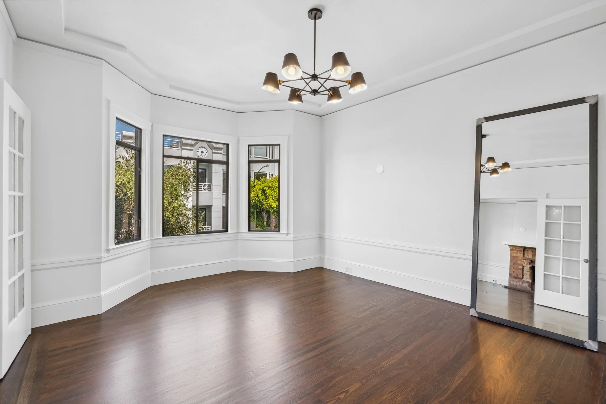 Empty room with wood flooring, large windows showing trees outside, a large mirror leaning against the wall, and a modern chandelier hanging from the ceiling.