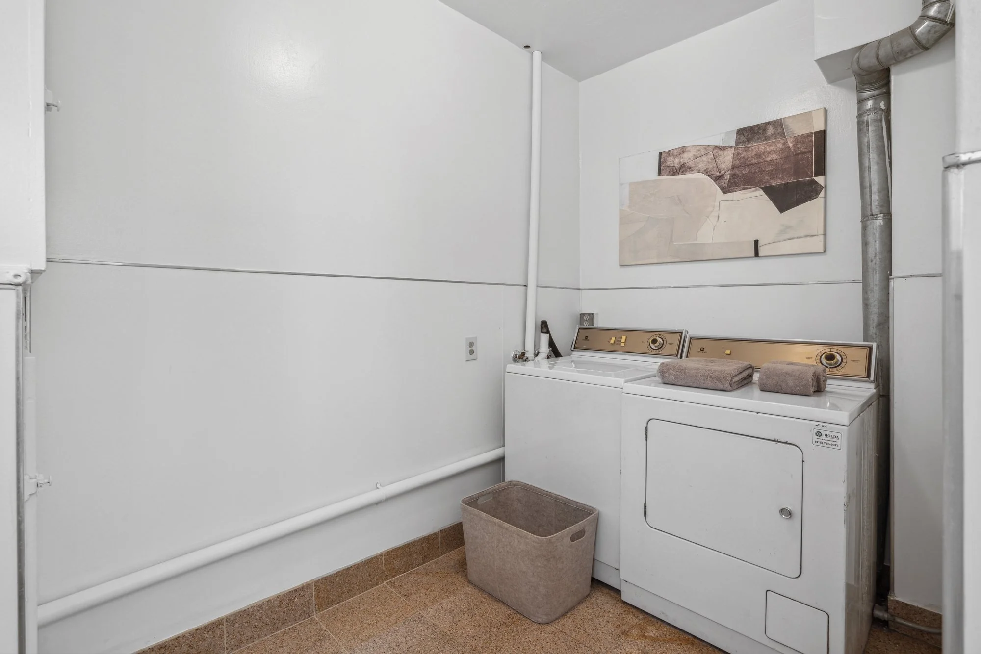 A laundry room with a white washing machine and dryer, beige tile floor, brown laundry baskets, and abstract artwork on the wall.