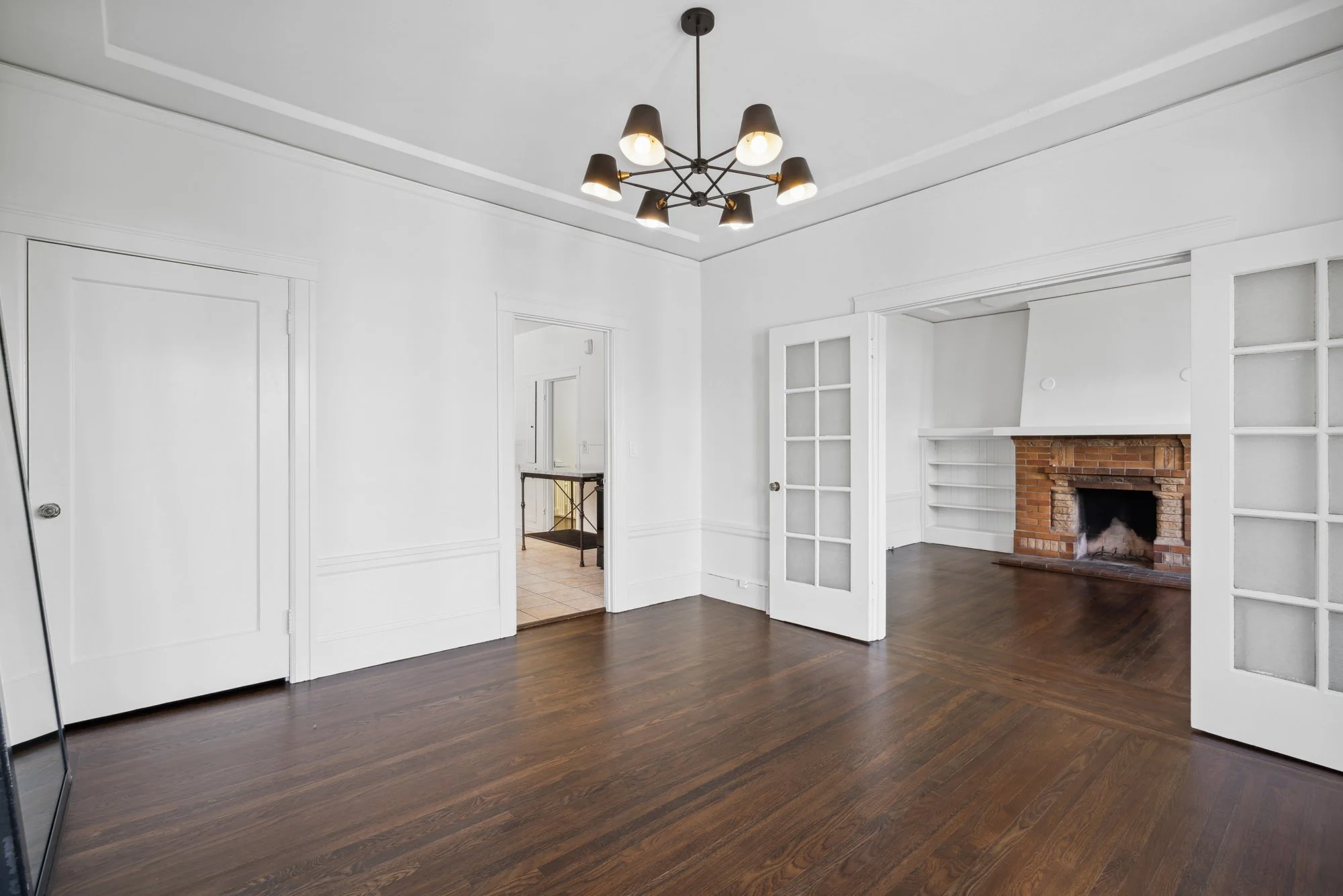 Empty living room with white walls, a brick fireplace, hardwood floors, and a modern chandelier
