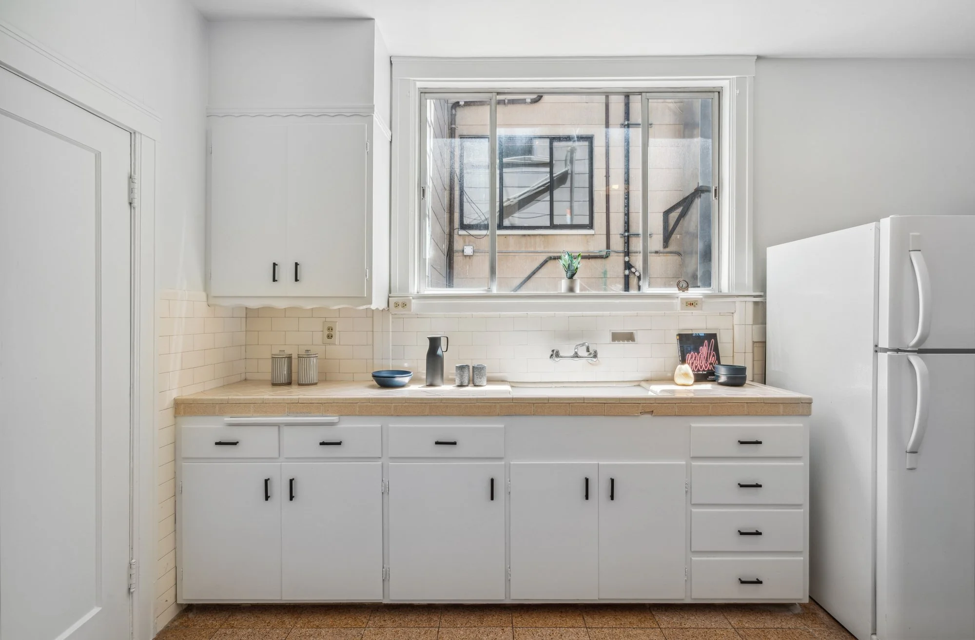 A minimalist kitchen with white cabinets, beige countertop, and a large window above the sink. The counter has various small decorative items and a small plant, with a fridge partially visible on the right.