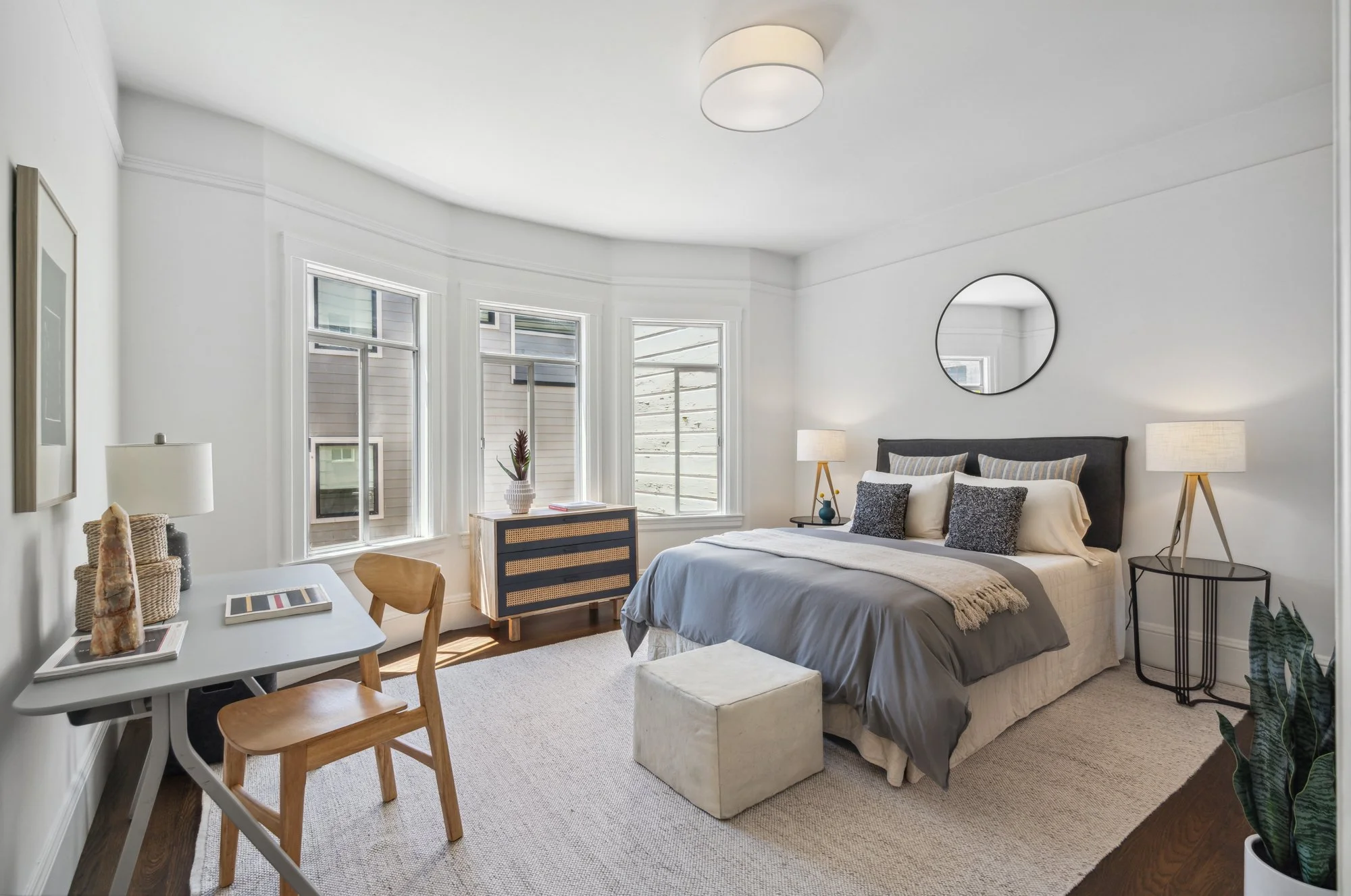 Bright bedroom with white walls, multiple windows, and a bed with white and gray bedding. Includes nightstands with lamps, a round mirror above the bed, and a small table with chairs and decorative items.