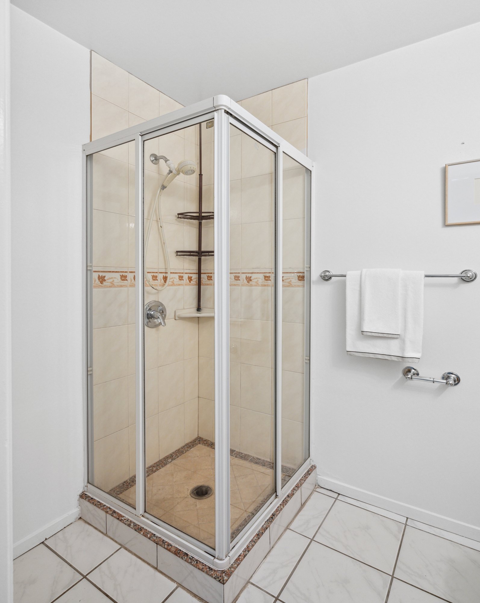 A small glass-enclosed shower with beige tile walls and a metal shower head inside. The shower is on a raised platform with a tiled border. To the right, a white towel hangs on a metal towel rack on a white wall. The floor outside the shower has whit
